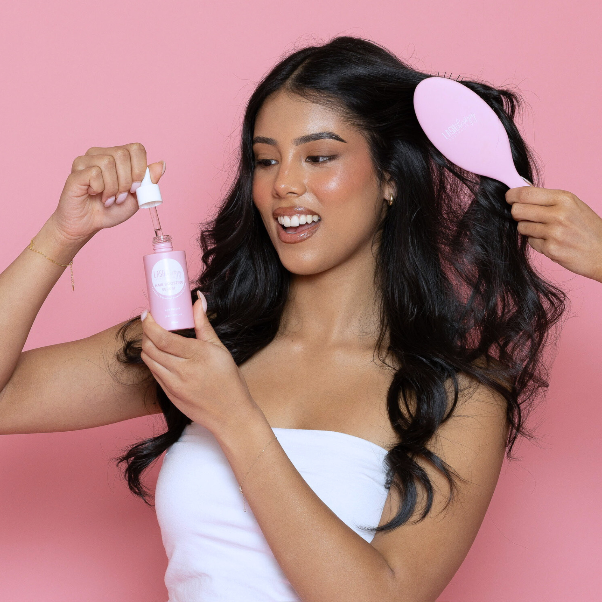 Woman with long dark hair holding a Lash Therapy Hair Boosting Peptide serum and Pink Hairbrush against a pink background